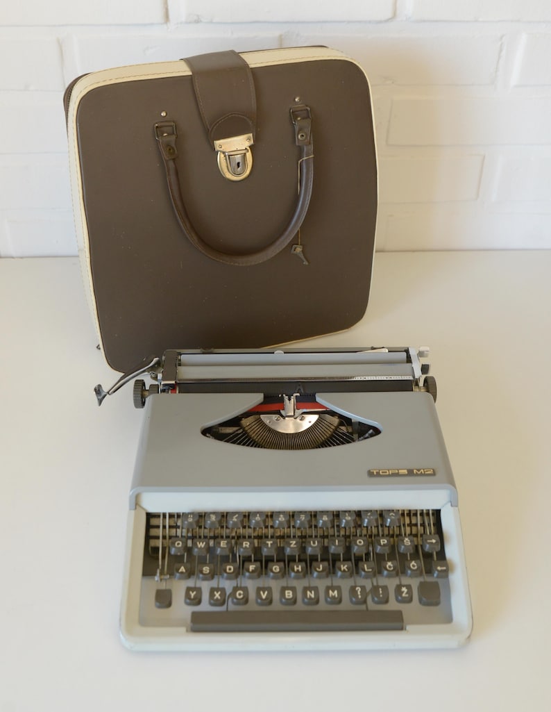 May include: A vintage gray TOPS M2 typewriter with a black keyboard and a brown carrying case. The typewriter has a silver carriage and a red ribbon indicator. The case is brown with a cream-colored trim and a handle.