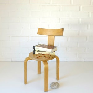 Può includere: Una sedia in legno con schienale curvo e quattro gambe. La sedia ha una seduta rotonda con due libri e un paio di occhiali sopra. La sedia è davanti a un muro di mattoni bianchi.