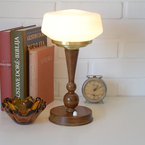 May include: A table lamp with a wooden base and a white glass shade, featuring a small white power switch on the circular base. The lamp is positioned next to three books, a small amber glass bowl with a ruffled edge, and a round metal alarm clock.