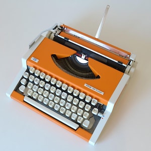 May include: A portable typewriter with a bright orange metal body and white frame. The keyboard features white circular keys with black lettering, including special characters. The brand name "ibm de Luxe" is printed on the front right side.