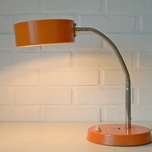 Puede incluir: L&aacute;mpara de escritorio naranja y plateada con un brazo flexible y una pantalla redonda. La l&aacute;mpara tiene un interruptor en la base.