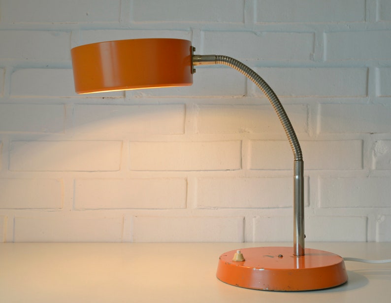Puede incluir: L&aacute;mpara de escritorio naranja y plateada con un brazo flexible y una pantalla redonda. La l&aacute;mpara tiene un interruptor en la base.