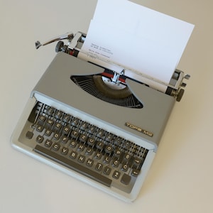 May include: A vintage gray TOPS M2 typewriter with a white sheet of paper inserted. The typewriter has a full keyboard with black keys and a silver carriage return lever. The paper is positioned to receive typed text.