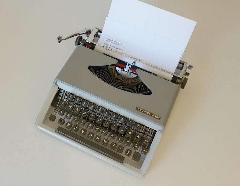 May include: A vintage gray TOPS M2 typewriter with a white sheet of paper inserted. The typewriter has a full keyboard with black keys and a silver carriage return lever. The paper is positioned to receive typed text.