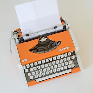 Könnte beinhalten: Eine leuchtend orangefarbene IBM de Luxe Schreibmaschine mit weißer Tastatur und einem Blatt Papier im Wagen. Das Gerät verfügt über ein metallisches Typensegment, einen silbernen Zeilenschalthebel und weiße Tasten mit schwarzer Beschriftung.