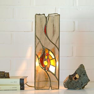 May include: A stained glass lamp made from an old book with a red, orange and yellow design. The lamp is lit and casting a warm glow.
