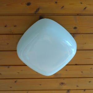 May include: A white, square-shaped light fixture with rounded corners. The fixture is mounted on a wooden surface with horizontal planks. The light has a glossy finish and reflects the surrounding light.