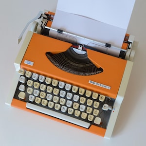 Op de afbeelding: Een oranje IBM de Luxe 1 schrijfmachine met een wit toetsenbord en toetsen. De schrijfmachine heeft een zwart lint en een vel wit papier erin. De schrijfmachine staat op een wit oppervlak.