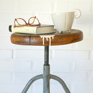 Könnte beinhalten: Ein vintage industrieller Hocker mit einer Holzplatte und einem grauen Metallgestell. Die Sitzfläche hat einen weißen Farbtropfen und ein Buch und eine weiße Keramiktasse stehen darauf.