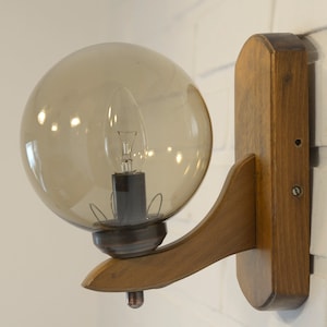 Puede incluir: Una lámpara de pared de madera marrón con una pantalla de vidrio transparente en forma de globo. La lámpara tiene un brazo curvo y un diseño sencillo.