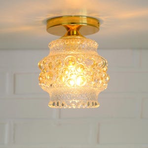 Pode incluir: Uma luminária de teto vintage com uma cúpula de vidro transparente texturizado e uma base de metal dourado. A cúpula tem um design semelhante a bolhas, e a luz está acesa, iluminando a luminária. A luminária está montada em um teto branco.