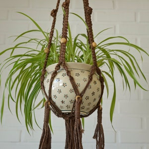 Puede incluir: Un macetero de macramé marrón con cuentas de madera sostiene una maceta de cerámica blanca con un patrón floral. La maceta está llena de una planta verde con hojas largas y delgadas.