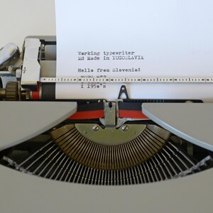 May include: Close-up of a vintage typewriter with a sheet of paper inserted. The paper displays text that reads "Working typewriter, Made in Yugoslavia, Hello from Slovenia! i 1950's". The typewriter is gray with black and red accents.