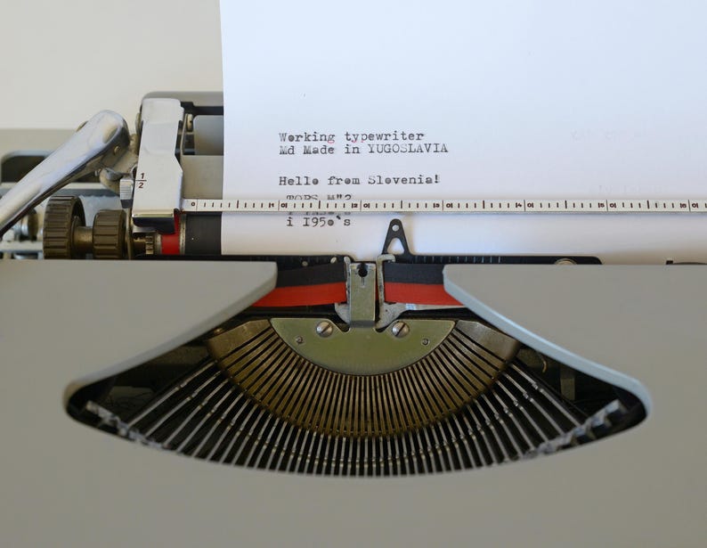 May include: Close-up of a vintage typewriter with a sheet of paper inserted. The paper displays text that reads "Working typewriter, Made in Yugoslavia, Hello from Slovenia! i 1950's". The typewriter is gray with black and red accents.