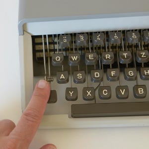 May include: Close-up of a vintage typewriter keyboard with gray keys and a gray frame. The keys are arranged in a QWERTY layout. A finger is pressing the 'A' key. The typewriter is on a white surface.