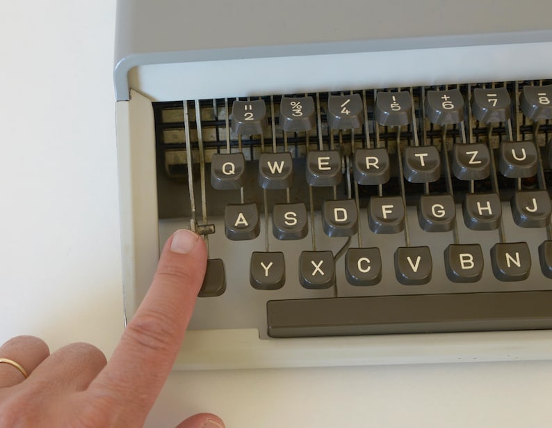 May include: Close-up of a vintage typewriter keyboard with gray keys and a gray frame. The keys are arranged in a QWERTY layout. A finger is pressing the 'A' key. The typewriter is on a white surface.
