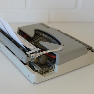 May include: A vintage gray and white typewriter with a sheet of white paper inserted. The typewriter has a silver carriage return lever and black keys. The machine is set on a white surface.