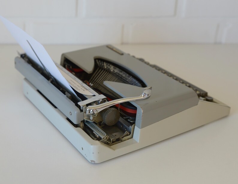May include: A vintage gray and white typewriter with a sheet of white paper inserted. The typewriter has a silver carriage return lever and black keys. The machine is set on a white surface.