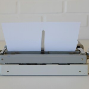 May include: A vintage typewriter with a white and gray body, holding a blank white sheet of paper. The typewriter has a classic design with silver metal accents and a lever on the right side. The paper is positioned in the center, ready for typing.