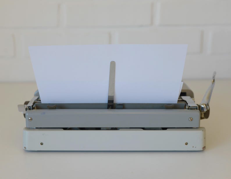 May include: A vintage typewriter with a white and gray body, holding a blank white sheet of paper. The typewriter has a classic design with silver metal accents and a lever on the right side. The paper is positioned in the center, ready for typing.
