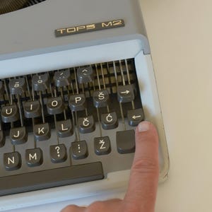 May include: Close-up of a gray TOPS M2 typewriter with a finger pressing the backspace key. The keyboard has gray keys with white lettering and symbols. The typewriter is on a white surface.