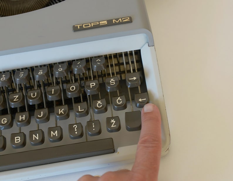 May include: Close-up of a gray TOPS M2 typewriter with a finger pressing the backspace key. The keyboard has gray keys with white lettering and symbols. The typewriter is on a white surface.