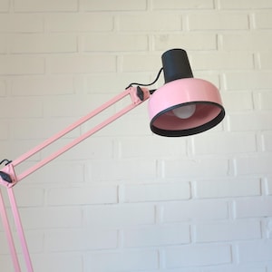 May include: A pink adjustable desk lamp with a black lampshade and a white lightbulb. The lamp has a long, articulated arm and a round lampshade. The lamp is set against a white brick wall.