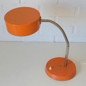 Puede incluir: L&aacute;mpara de escritorio naranja con un brazo flexible y una pantalla redonda. La l&aacute;mpara tiene un cable blanco y un peque&ntilde;o interruptor en la base.