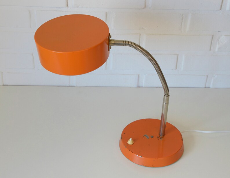 Puede incluir: L&aacute;mpara de escritorio naranja con un brazo flexible y una pantalla redonda. La l&aacute;mpara tiene un cable blanco y un peque&ntilde;o interruptor en la base.
