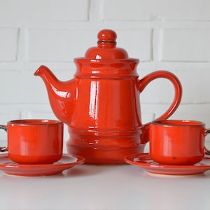 May include: A red ceramic teapot with a lid and two matching teacups and saucers.