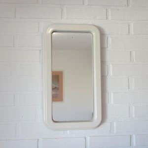 May include: A rectangular mirror with rounded corners, framed in a cream-coloured border. The mirror is mounted on a white brick wall. The reflection shows a glimpse of a framed picture. The mirror is a decorative item.