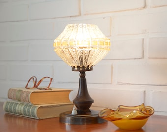 Può includere: Una lampada da tavolo vintage con un paralume in vetro trasparente con un bordo dorato. La lampada ha una base in ottone ed è accesa su un tavolo in legno con una pila di libri e un piccolo posacenere in vetro ambrato.