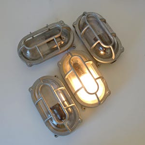 May include: Four oval-shaped, silver-colored outdoor wall lights. Two lights are illuminated with a warm glow, while the other two are unlit. Each light has a protective metal cage and a clear glass cover.