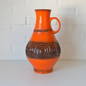 May include: Orange ceramic vase with a brown swirl design and a handle. The vase has a textured surface.