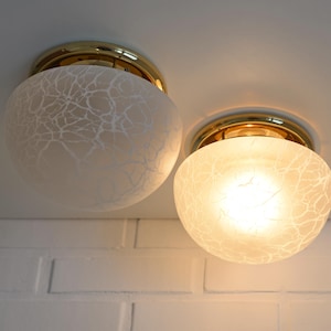 May include: Two ceiling lights with gold-coloured trim. The frosted glass globes have a white, crackled pattern. One light is illuminated, casting a warm glow. The fixtures are mounted on a white ceiling above a white brick wall.