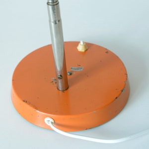 Puede incluir: Base de metal naranja de una l&aacute;mpara de mesa con un v&aacute;stago de metal plateado y un cable blanco.