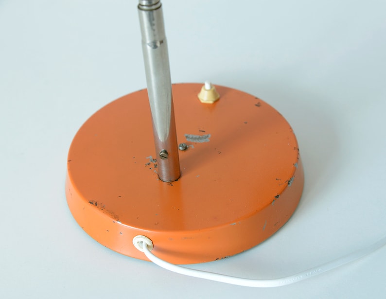 Puede incluir: Base de metal naranja de una l&aacute;mpara de mesa con un v&aacute;stago de metal plateado y un cable blanco.