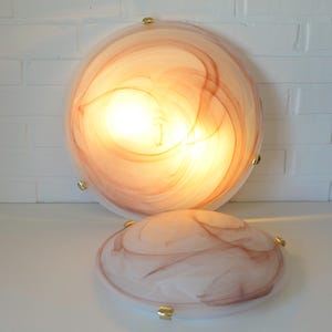 May include: Two round, flush-mount ceiling lights with a swirling, marbled design in shades of peach and brown. The lights have a brass-colored trim and are illuminated, casting a warm glow. One light is larger than the other.