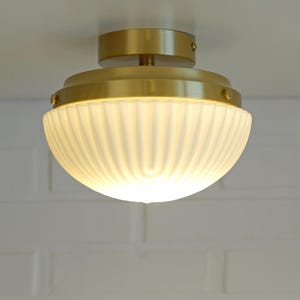 Può includere: Un lampada da soffitto con finitura in ottone e paralume in vetro bianco a coste. L'apparecchio è rotondo ed emette una luce soffusa e calda. Il design è in stile classico e vintage, adatto all'illuminazione domestica.