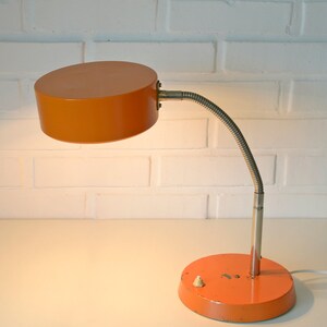 Puede incluir: Una l&aacute;mpara de escritorio naranja con un brazo flexible y una pantalla redonda. La l&aacute;mpara tiene un cable blanco y un peque&ntilde;o interruptor en la base.