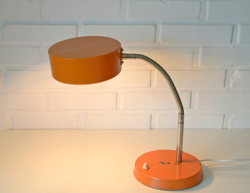 Puede incluir: Una l&aacute;mpara de escritorio naranja con un brazo flexible y una pantalla redonda. La l&aacute;mpara tiene un cable blanco y un peque&ntilde;o interruptor en la base.