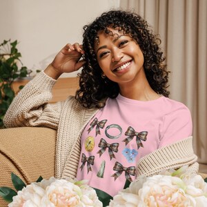 May include: A light pink short-sleeved t-shirt featuring a repeating pattern of brown plaid bows, a small silver ring, a yellow circular object, a green vertical stone, and a cluster of small light blue flowers printed on the front.