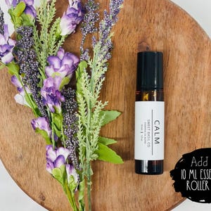 Puede incluir: Un frasco de aceite esencial de 10 ml con un rodillo, tapa negra y una etiqueta blanca que dice "CALM SWEET BASIL CO" y "ENDING" en texto negro. El frasco est&aacute; sobre una superficie de madera con flores moradas.
