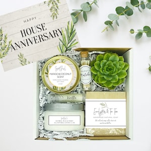 May include: A white gift box filled with a housewarming gift set. The set includes a mango and coconut scented candle, a vanilla scented candle, a small succulent plant, a bar of eucalyptus and tea tree soap, and a small bottle of safety matches. The box is decorated with a card that says "Happy House Anniversary".