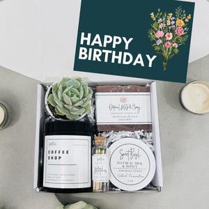 May include: A gift box filled with a small succulent plant, a candle, a bar of soap, and a small jar of lotion. The box is decorated with shredded paper and a card that says "Happy Birthday".