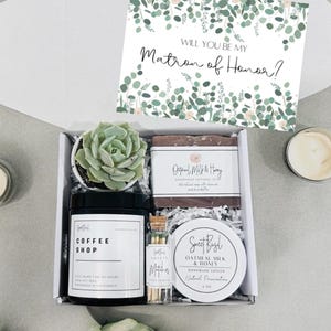 May include: A gift box with a card that says "Will you be my Matron of Honor?" The box contains a small succulent plant, a candle, a bar of soap, and a small jar of lotion.