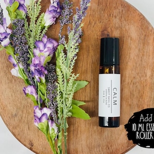 Może przedstawiać: 10 ml butelka z olejkiem eterycznym z rolką, czarną zakrętką i białą etykietą z napisem "CALM" i "SWEET BASIL CO". Butelka znajduje się na drewnianej powierzchni z fioletowymi kwiatami.