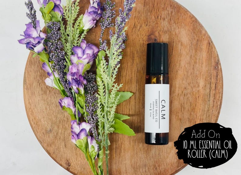 Może przedstawiać: 10 ml butelka z olejkiem eterycznym z rolką, czarną zakrętką i białą etykietą z napisem "CALM" i "SWEET BASIL CO". Butelka znajduje się na drewnianej powierzchni z fioletowymi kwiatami.