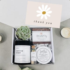 May include: A gift box filled with a small succulent plant, a candle, a bar of soap, and a small jar of lotion. The box is lined with white tissue paper and has a card that says "thank you".