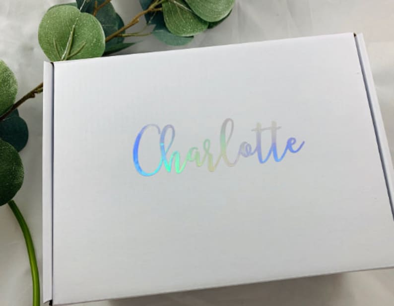 Może przedstawiać: Białe pudełko kartonowe z nadrukiem imienia "Charlotte" w iryzujących literach.